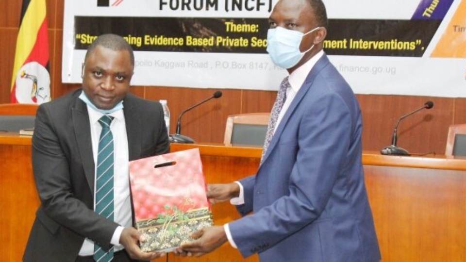 Two officials in face masks exchanging a token of appreciation in front of the 11th National Competitiveness Forum backdrop, which carries the Forum theme.